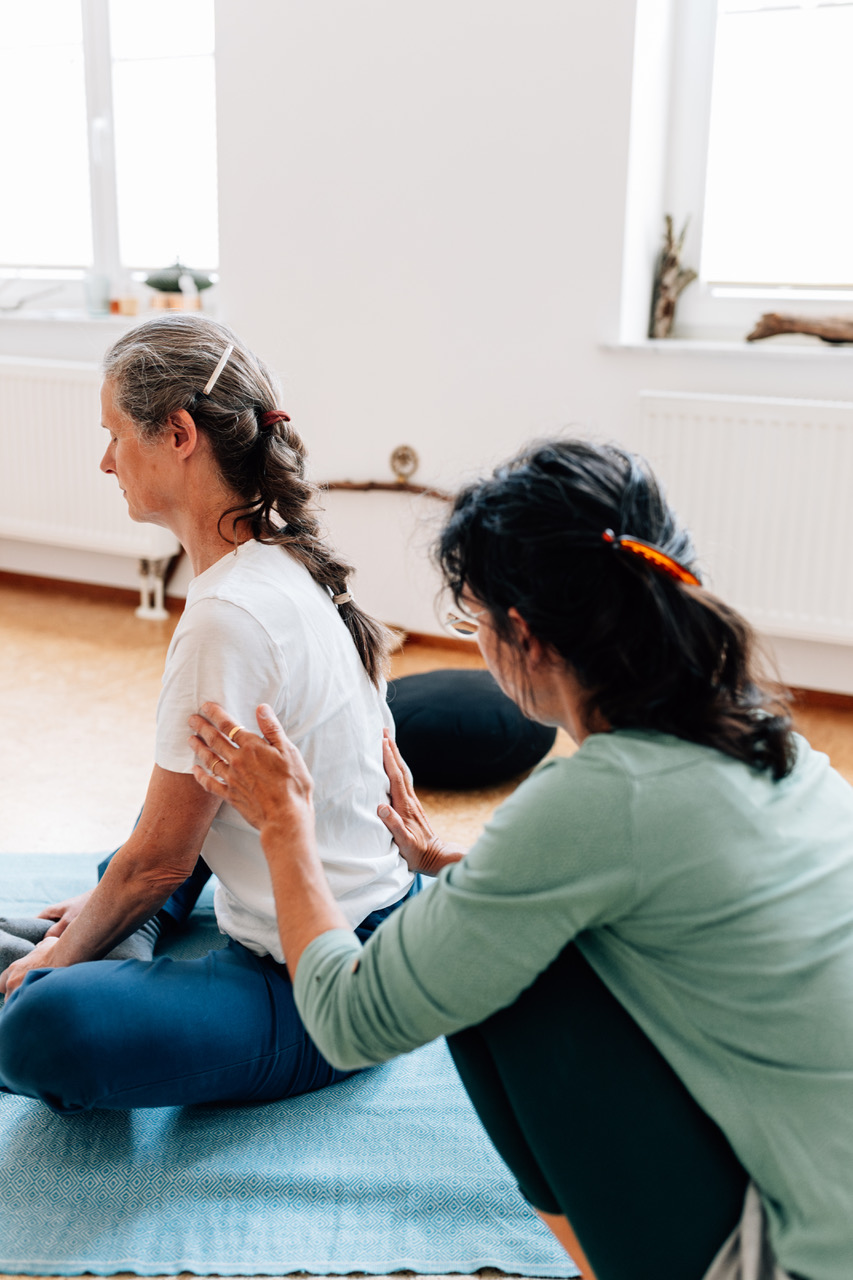 Die Yogalehrerin Maria Lichtenberg korrigiert eine Yogaschülerin in einer Variante der Schildkröte (Kurmasana Var.). Es geht um die Verlängerung der Wirbelsäule von der Wurzel beginnend. Das Bild ist von seitlich hinten aufgenommen.