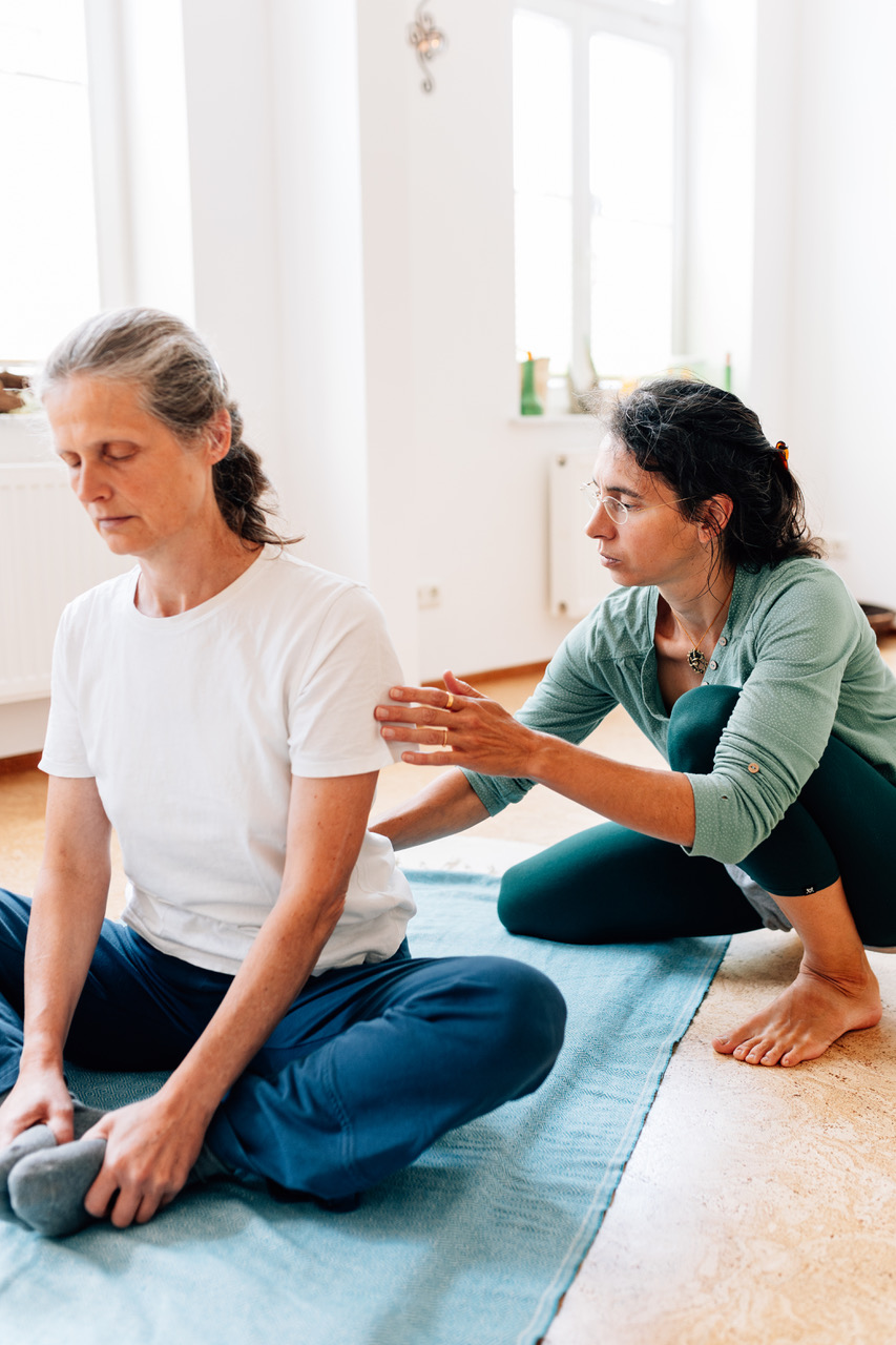 Die Yogalehrerin Maria Lichtenberg korrigiert eine Yogaschülerin in einer Variante der Schildkröte (Kurmasana Var.). Es geht um die Verlängerung der Wirbelsäule von unten beginnend. Das Bild ist von seitlich vorn aufgenommen.